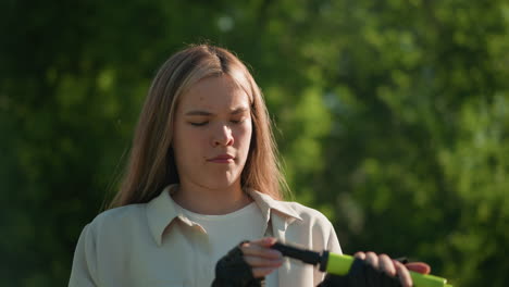 chic blanca con cabello rubio examina el mango de la bomba de aire verde con una expresión de disgusto, mostrando su reacción en un entorno natural al aire libre con árboles en el fondo