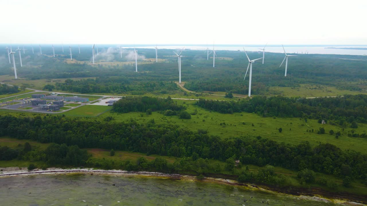 Aerial drone footage of a large misty and foggy peninsula at a beach seaside shoreline where large white wind turbines are generating renewablr energy power during misty and cloudy day in Paldiski