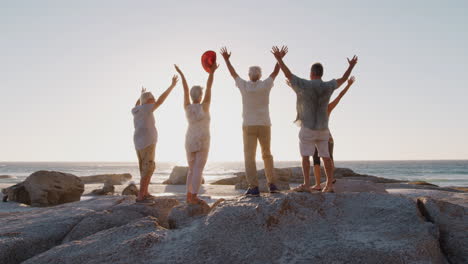 Rückansicht-älterer-Freunde,-Die-Im-Gruppenurlaub-Auf-Felsen-Stehen-Und-Die-Arme-Heben-Und-Auf-Das-Meer-Blicken