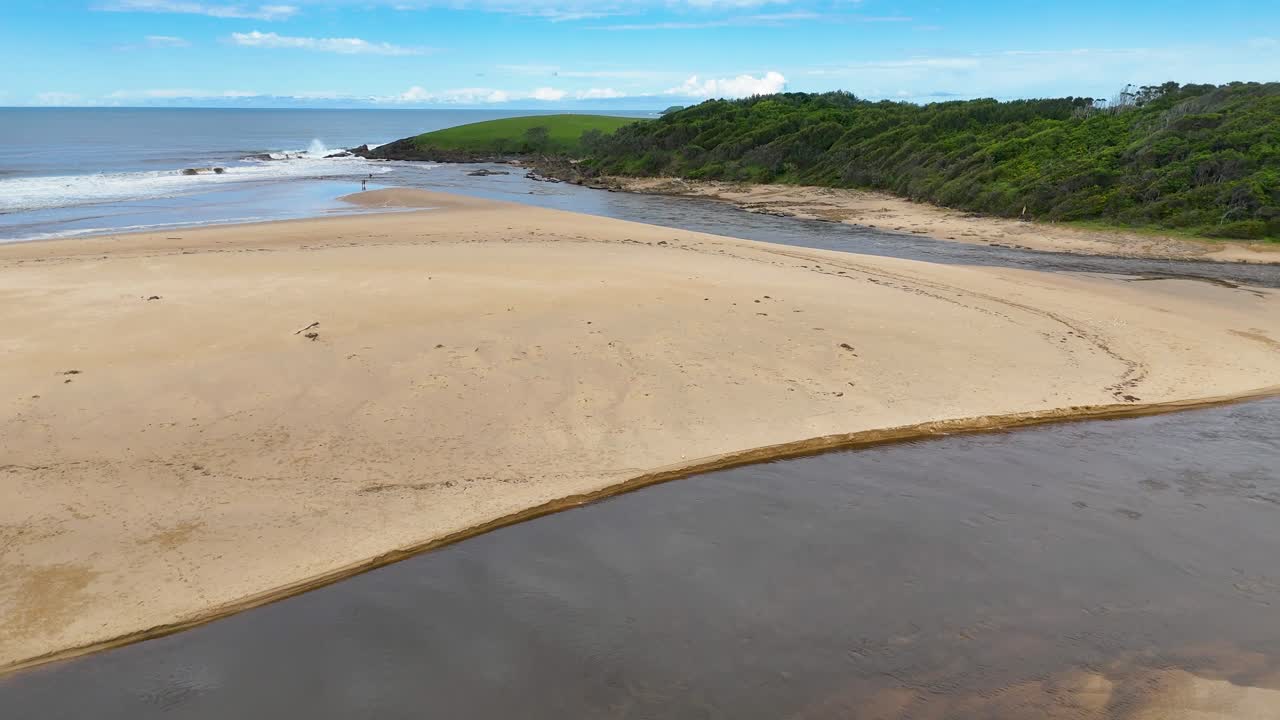 Drone pans across sandy estuary, tidal river, and ocean under bright daylight at Moonee Beach