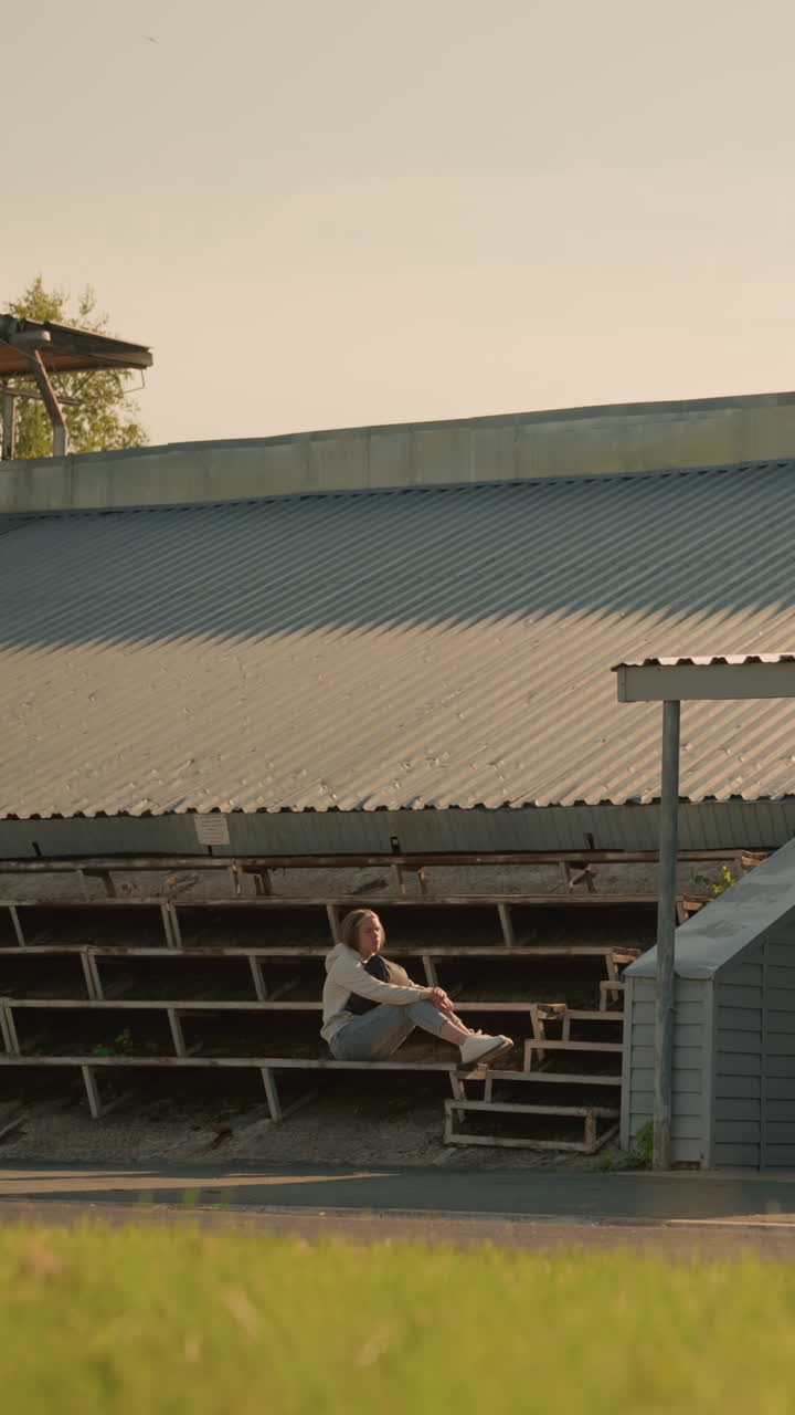 스테디움의 관중석에 혼자 앉아있는 여성, 반사 포즈, 무에 손을 고, 배경에는 자연 나무와 초록색으로 된 금속 스타디움 지붕 구조가 포함되어 있습니다.