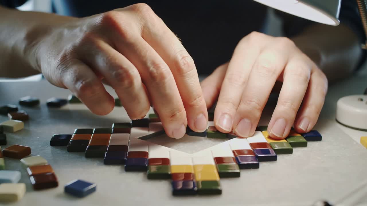 Manos suspendidas recogiendo teselas de mosaico, colocándolas para rellenar una plantilla de diamante en una mesa de trabajo, con la lámpara encendida