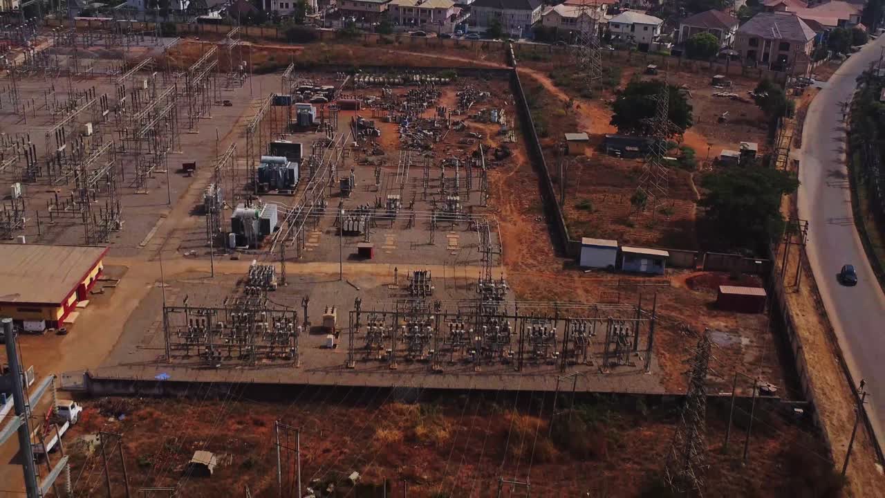 Beautiful aerial of a large power grid substation in Abuja, Nigeria on a sunny day. The drone flies backwards past a large electricity pylon and a car drives over a nearby road