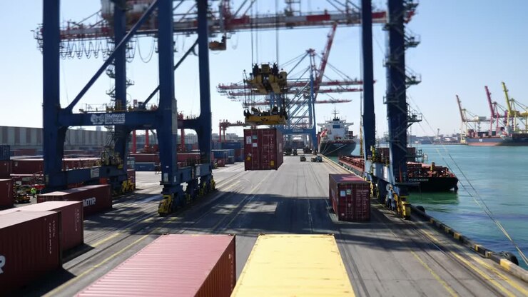 Heavy machinery is in action at a bustling port where large container cranes lift cargo from trucks. Containers are lined up along the dock while ships await loading operations.