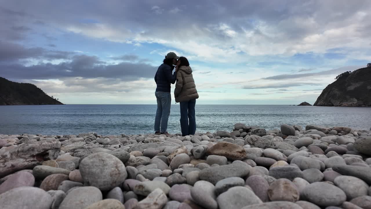 A couple kisses on a rocky shoreline facing the sea under a dramatic cloudy sky