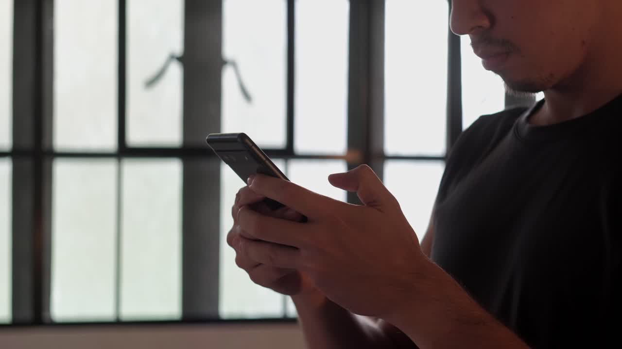 Medium shot of a young man using modern smartphone indoors while standing near a window with natural light
