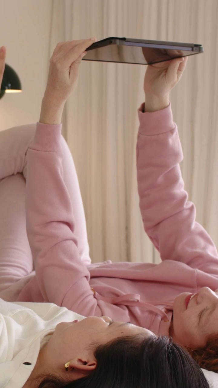 Vertical video: Swiping two women sharing tablet, lying on bed, wearing white and pink sweatshirts