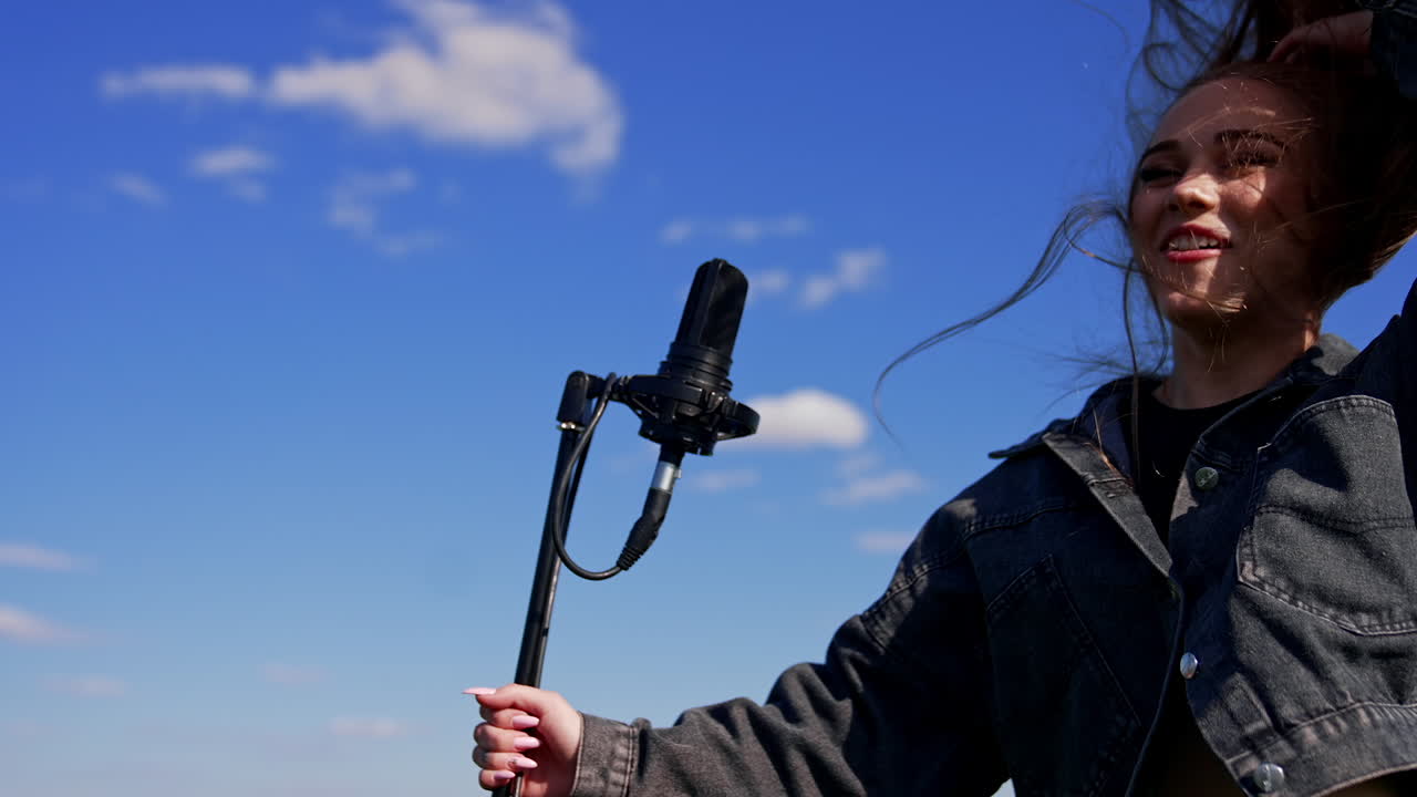 Happy girl singing and dancing in front of microphone outdoors. Female singer moving her head with fluttering hair on blue sky background.