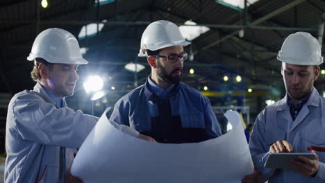 Drei-Ingenieure-Mit-Helmen-Halten-Bauplan-Und-Tablet-In-Der-Hand,-Während-Sie-Sich-In-Einer-Fabrik-Unterhalten