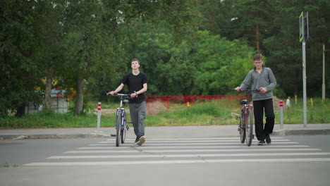 two friends walking side by side with their bicycles on a pedestrian crossing marked with white paint, the background features a car passing by, green trees, and a bright red fence