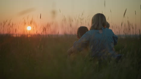 a back view of a mother in a blue gown sitting in a field with her two young children beside her. they are all watching the serene sunset over a tranquil lake, surrounded by tall grasses