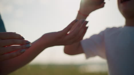 close-up of a woman's hands carefully cleaning a red apple before handing it to a child wearing a white shirt and glasses. the scene is set outdoors with a bright, natural background