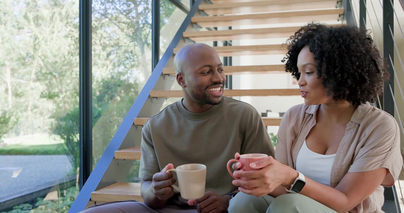 African American couple sitting on stairs near window, man starting chat while connecting with mugs