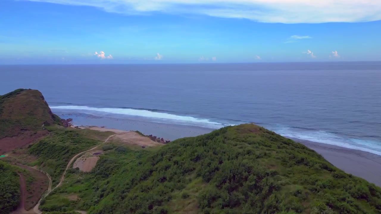 Lombok island coastline view showing lush green hills, a winding path, distant ocean waves, and clear shallow water. Wonderful aerial view speed ramp hyper motion time lapse overflight flyover