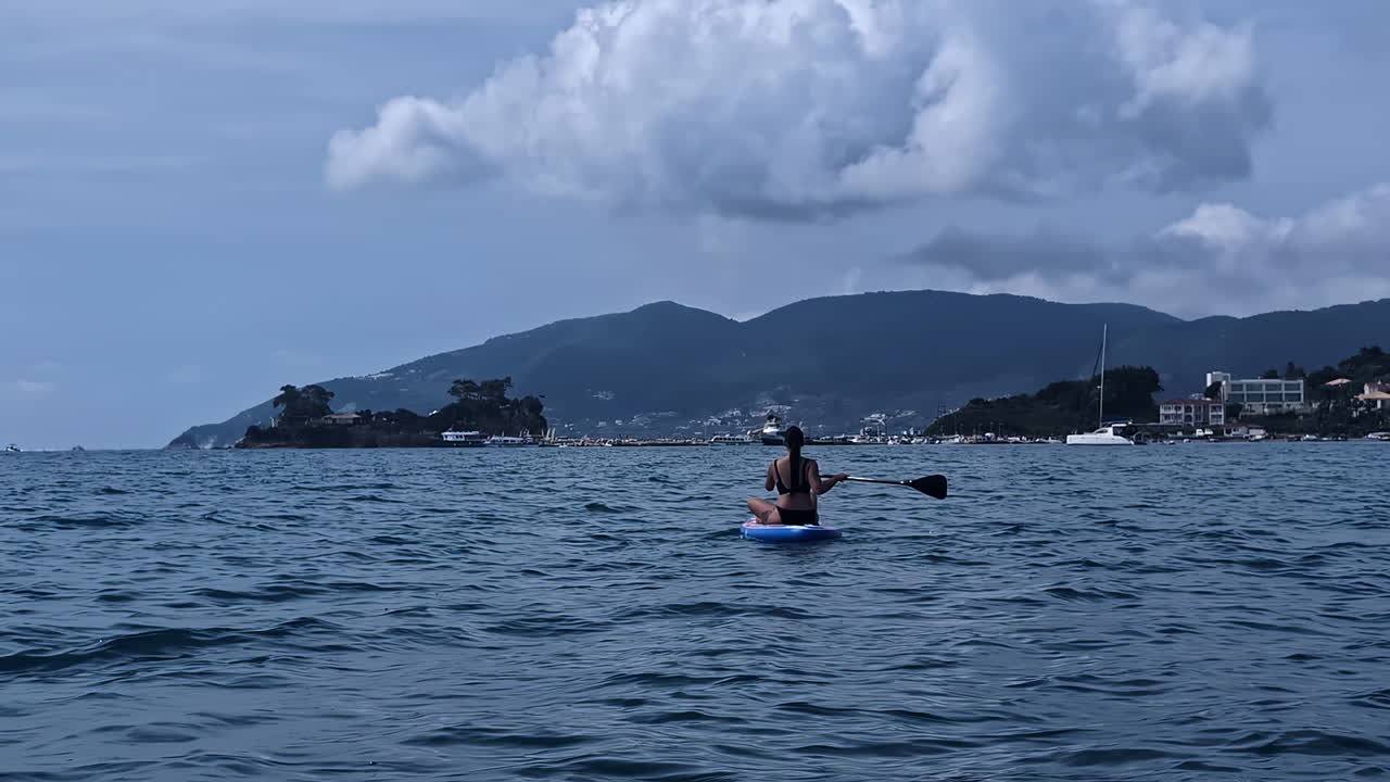 A white woman is paddling on a blue board while sitting and enjoying the view of the cloudy mountains, slowm motion and copy space