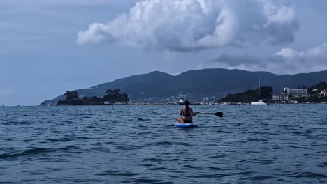 a white woman is paddling on a blue board while sitting and enjoying the view of the cloudy mountains, slowm motion and copy space