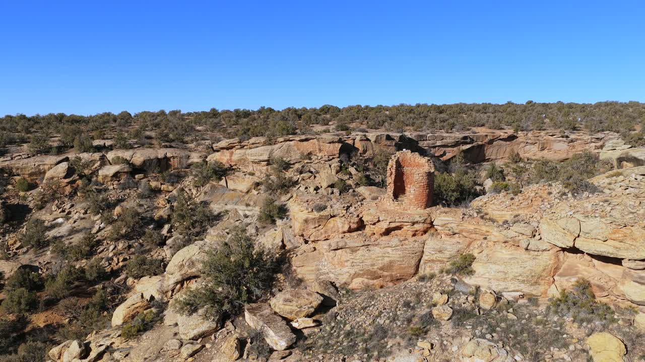 An ongoing aerial orbit captures an ancient masonry tower built onto a sheer cliff ledge. The shot emphasizes the structure's isolation within a rugged and vast pinyon-juniper woodland