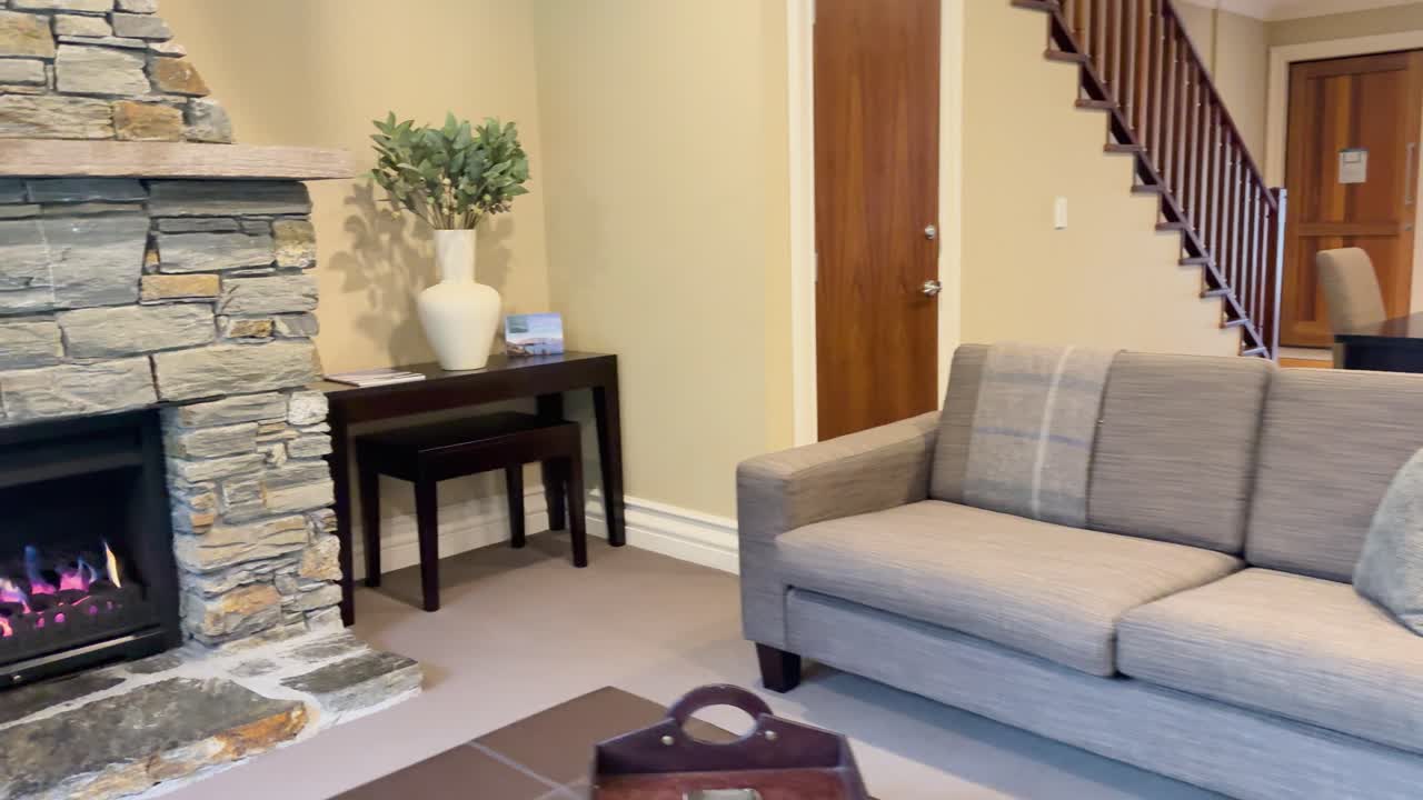 A bright, contemporary living room with beige sofas, stone fireplace, and wooden accents. Smooth camera pan highlights cozy seating, natural light, and elegant interior styling