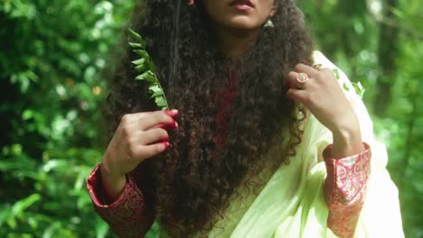 close-up of a woman in a green sari, adorned with henna and a flower in her hair, gracefully raising two leafy branches along her head