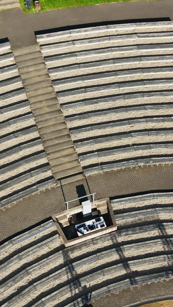 Sunlit Sun Hill open air amphitheater in Talsi, Latvia, displaying empty seats, control booth, and paved walkways from overhead perspective, capturing architectural serenity, vertical drone shot