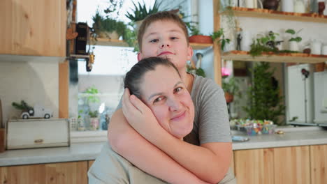 boy hugs mom from the back, both in gray clothes, smiling happily in kitchen setting, family love and happiness captured, joyful moment between mother and son, indoor family bonding
