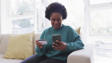 Happy-african-american-woman-making-payment-using-credit-card-and-smartphone-at-home,-slow-motion