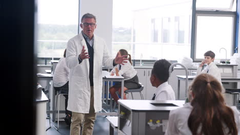 male high school tutor teaching students in uniforms sitting at work benches in science class