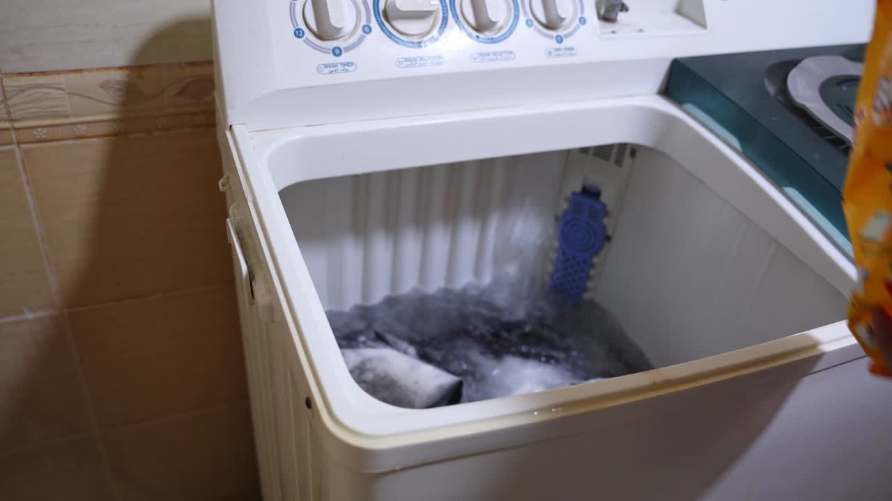 Close-up of white granular laundry detergent powder being poured into a top-loading washing machine filled with clothes. Authentic domestic laundry routine