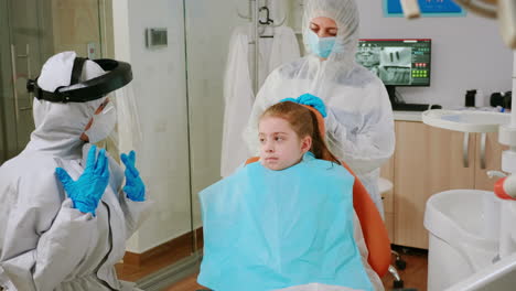 Little-girl-with-dental-bib-and-ppe-suit-listening-pediatric-dentist-in-coverall