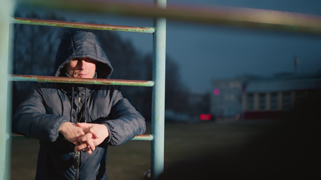 Close-up of a person in a hooded jacket resting on an iron bar with clasped hands, illuminated by light, the background features a blurred view of a yellow building and a moving car