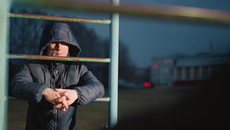 close-up of a person in a hooded jacket resting on an iron bar with clasped hands, illuminated by light, the background features a blurred view of a yellow building and a moving car