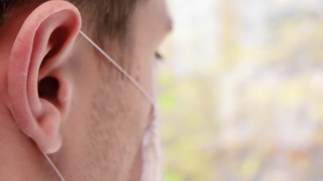 Caucasian man putting black wireless earbud in the ear while wearing an protective medical mask. Rear view. Close up. Quarantine and Corona Virus idea