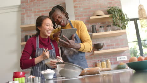 Happy-diverse-couple-in-aprons-using-tablet-and-baking-in-kitchen,-copy-space,-slow-motion