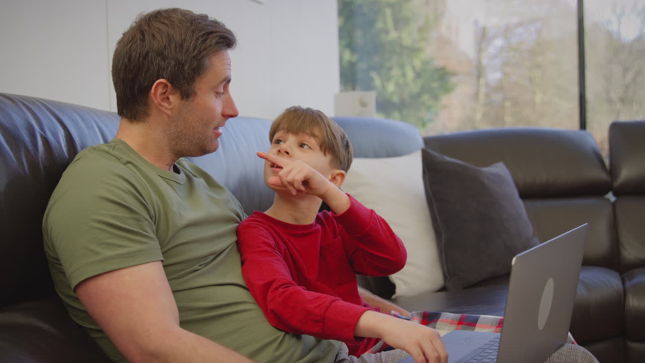 padre e hijo sentados en el sofá en casa en pijama juntos mirando la computadora portátil - filmado en cámara lenta