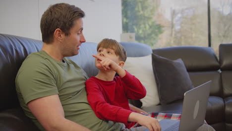 father and son sitting on sofa at home in pyjamas together looking at laptop - shot in slow motion