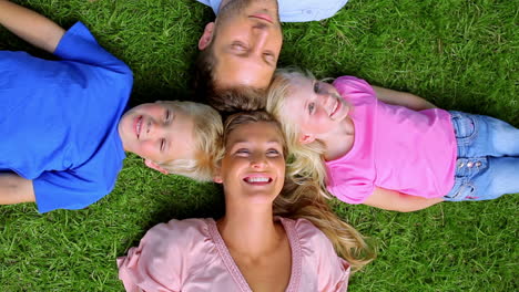 Toma-Cenital-De-Una-Familia-Sonriendo-Mientras-Yacen-Cabeza-Con-Cabeza-En-El-Césped.