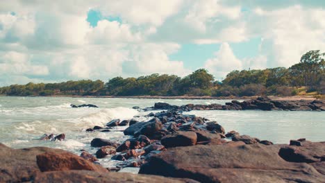 Small-waves-crash-against-a-natural-wall-made-of-small-rocks-whilst-fluffy-white-clouds-float-above-a-rocky-beach-and-bush-treeline-on-a-beautiful,-warm,-sunny-day