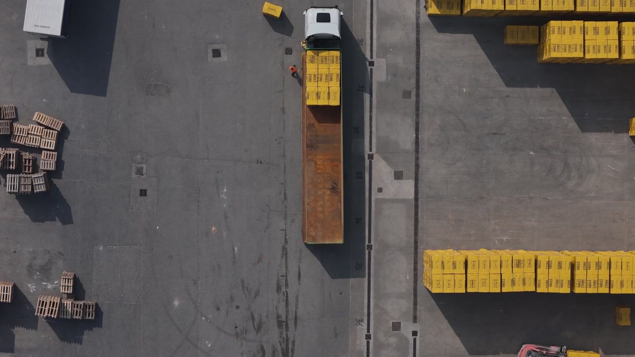 Top down drone tracking shot of forklift loading prefabricated concrete blocks onto transport truck inside industrial storage facility in Po Valley Italy for construction supply chain distribution
