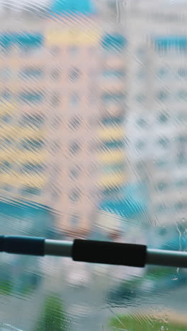 brush cleaning dirty window with water and soap. washing glass window inside on city building background. vertical video