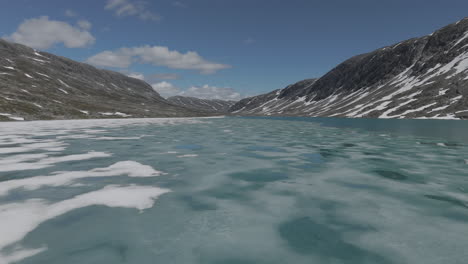 um drone em câmera lenta voando perto dos flocos de gelo no lago langvatnet, na noruega, perto do strynefjellsveg em um dia ensolarado, cercado por rochas brilhantes e montanhas.