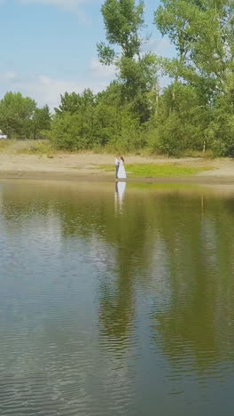 aerial motion above tranquil lake to newly wedded couple standing on green bank near green forest