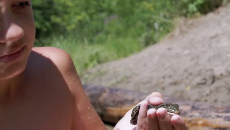 niño sostiene una rana verde en la mano contra el fondo de la orilla arenosa del río