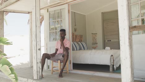 Thoughtful-african-american-man-sitting-on-porch-of-wooden-beach-house-looking-away,-slow-motion