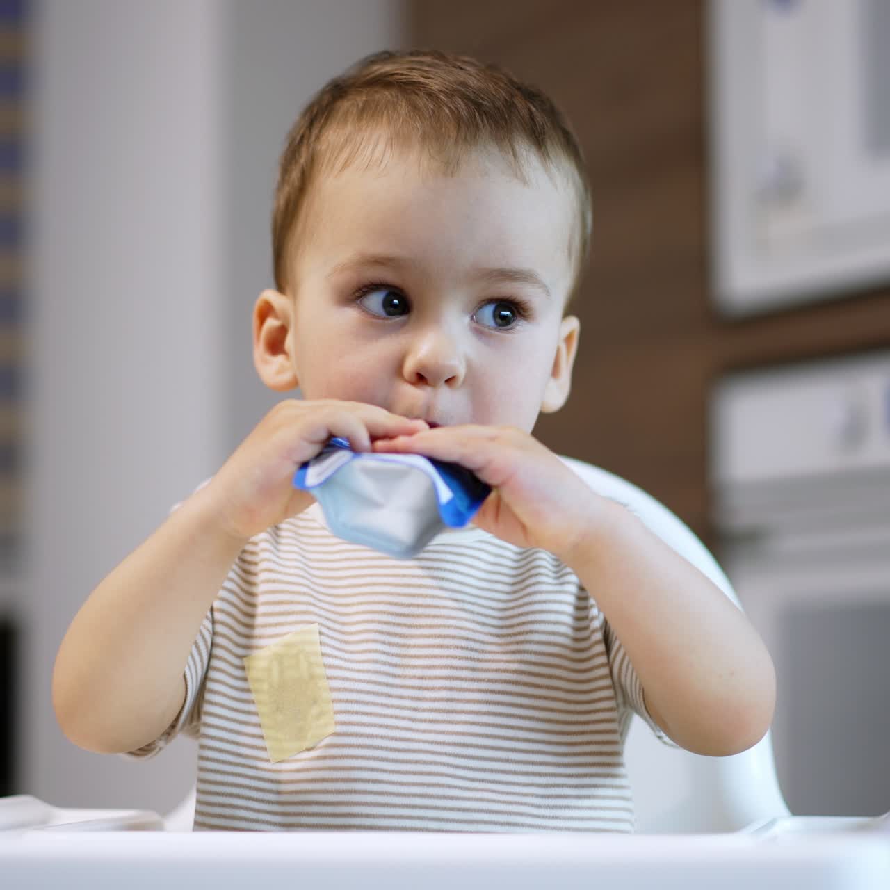 Adorable baby boy takes a pack of food into mouth. Little kid eating the pureed fruit willingly. Blurred backdrop