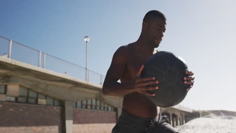 homme afro-américain concentré soulevant une balle, faisant de l'exercice à l'extérieur sur la plage