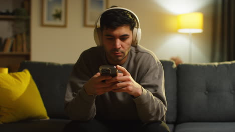 man spending evening at home sitting on sofa wearing wireless headphones streaming music or watching content from mobile phone