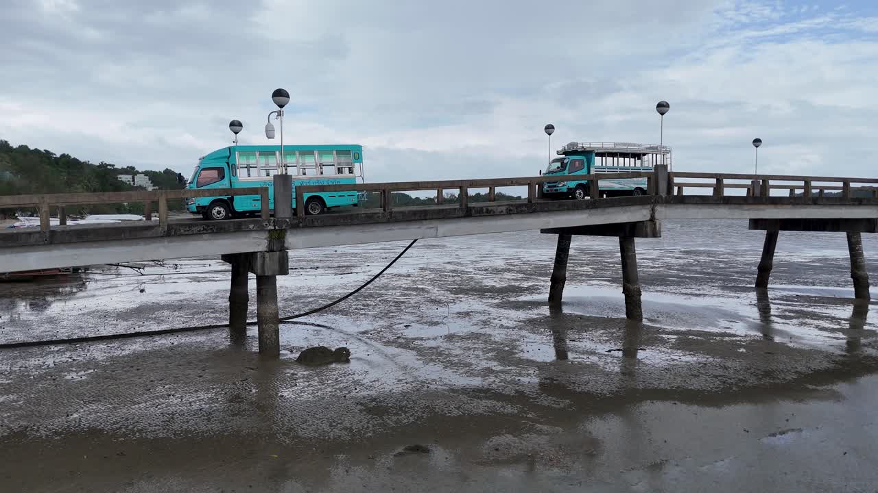 A turquoise bus navigates a narrow pier in Phuket, Thailand, under overcast skies, showcasing local transportation and scenic coastal views