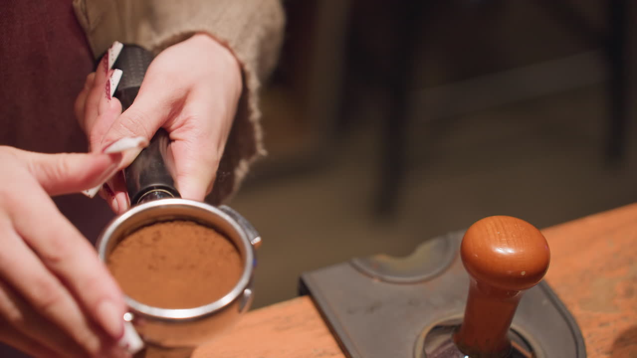 Close up of bartender using tamper to press coffee grounds into portafilter with focused hand movement over bar counter, warm lighting and soft textures