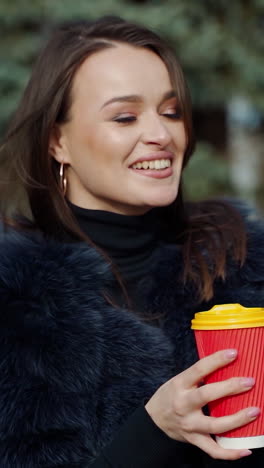 portrait of a happy girl in a sunny day in winter. beautiful woman in fur coat holds a cup of hot drink in the park and smiling at camera. slow motion. vertical video