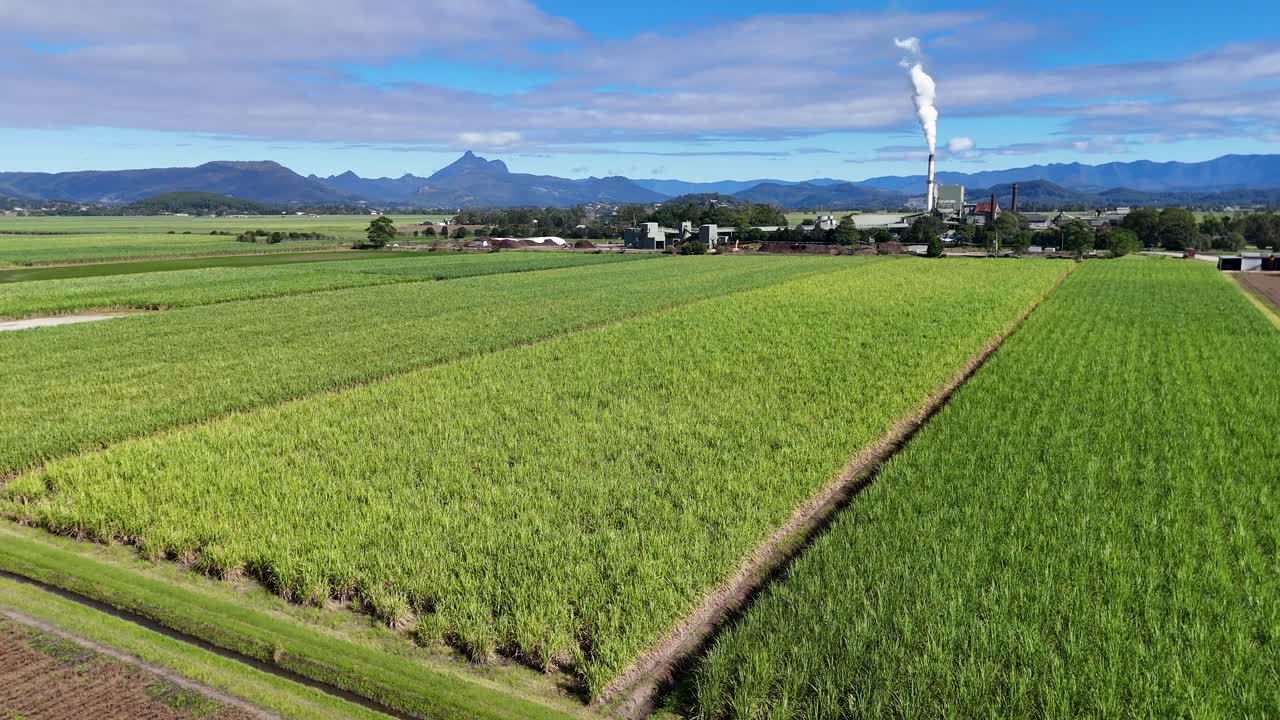 Drone footage over lush green fields and a distant factory emitting smoke under clear skies